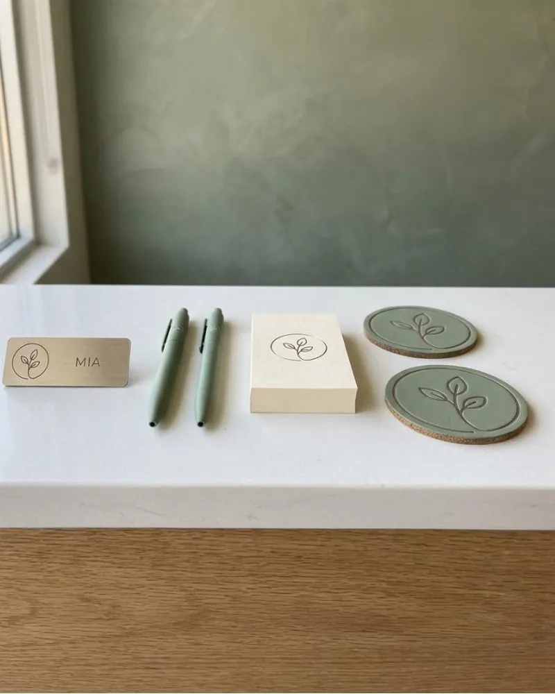 Branded front-desk pieces on a reception counter: a champagne-gold magnetic name tag, two sage pens, a stack of cream appointment cards and two sage coasters, each with the same mark.