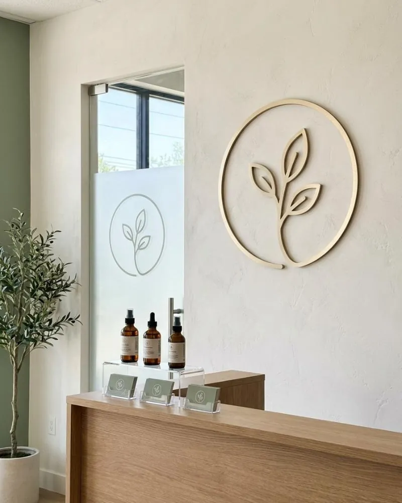 A med spa reception wall with a dimensional champagne-gold mark mounted above the desk, a frosted decal of the same mark on the glass door, and small shelf cards on a retail shelf.